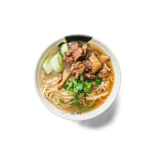 A bowl of noodle soup with sliced meat, herbs, and greens on a white background