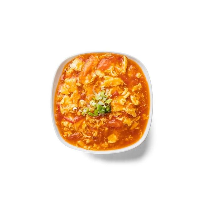 Square bowl of orange tomato soup topped with herbs on a white background