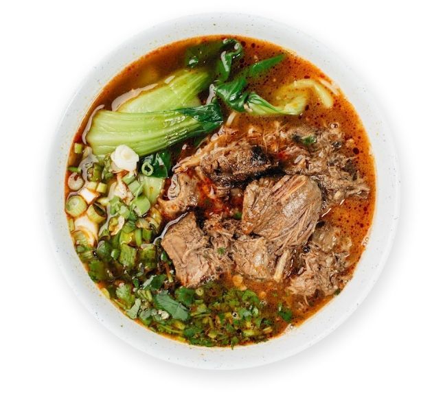 Bowl of beef noodle soup topped with herbs, greens, and chili oil