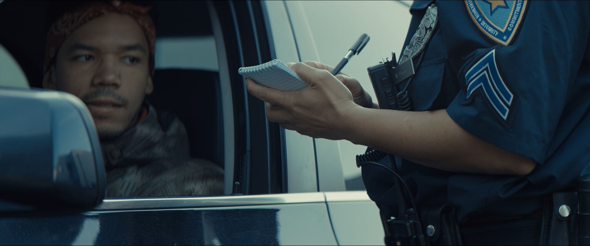 A police officer in uniform stands by the window of a silver sedan, handing a document to the driver on a suburban road. A police officer in uniform stands by the window of a silver sedan, handing a document to the driver on a suburban road.