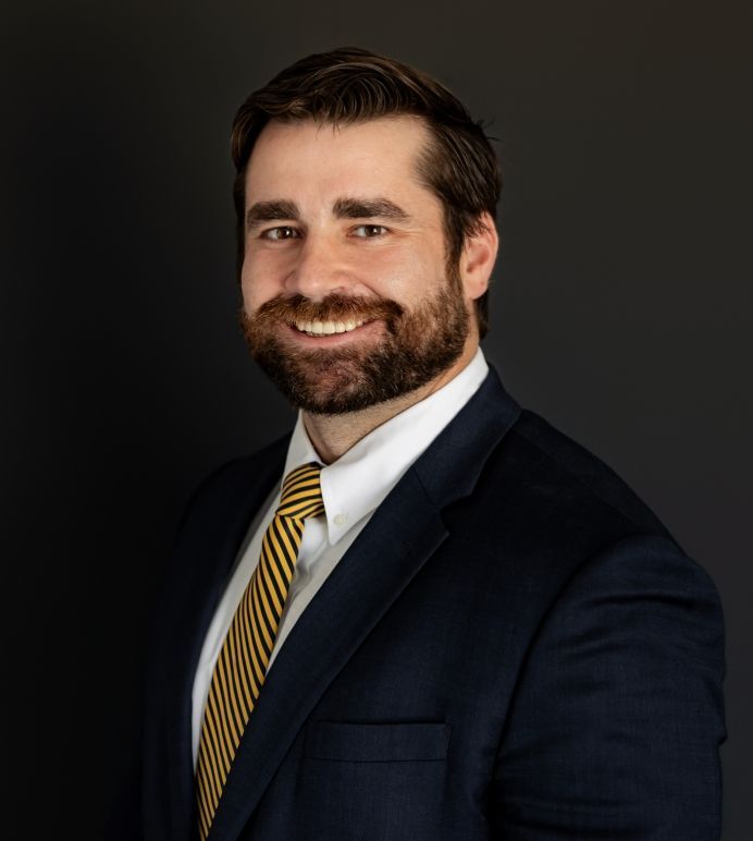 A professional in a navy suit and patterned tie sitting in an office chair in front of a bookshelf filled with books.