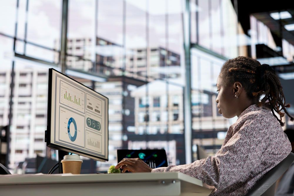 Person working at a desk with dual monitors in a bright office, reviewing charts and graphs