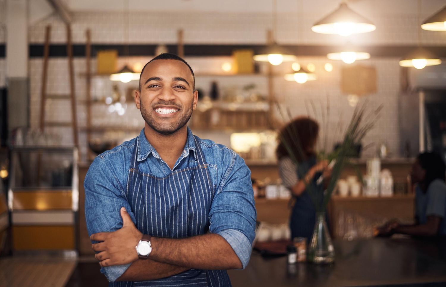 Smiling man in a blue shirt stands with arms crossed in a cozy café.