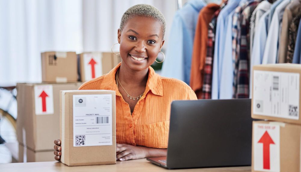 Person smiling at a laptop beside stacked shipping boxes in a small workspace
