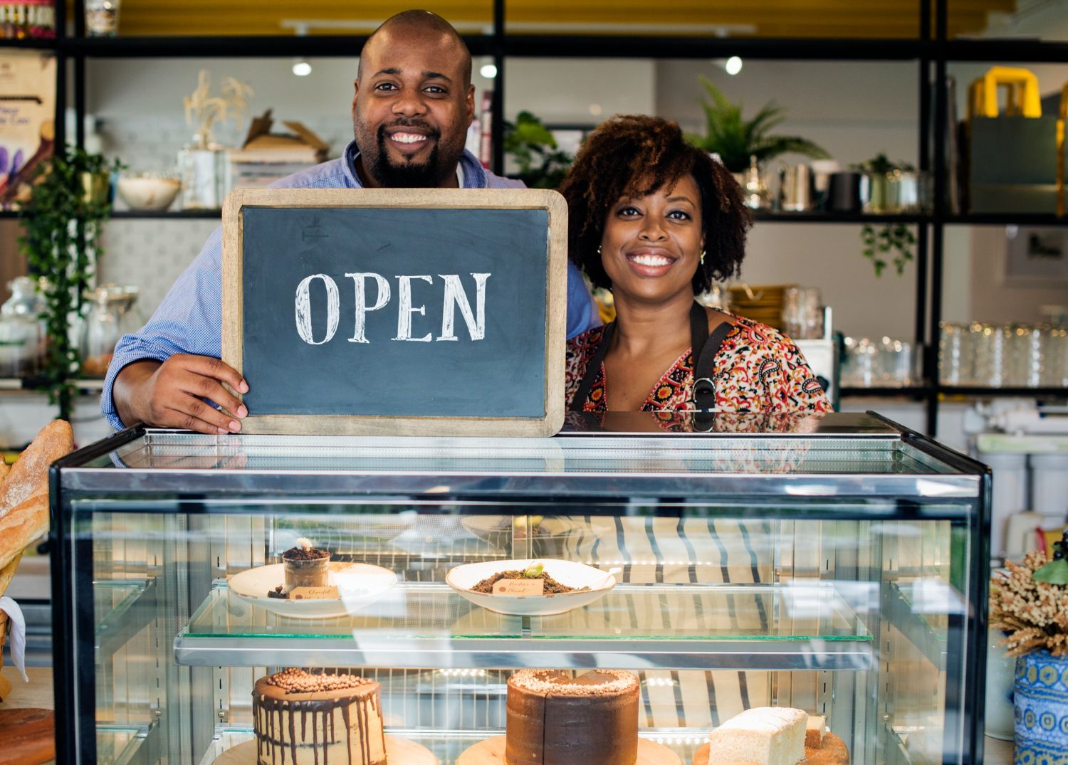 Two people smiling behind a bakery counter, holding an “OPEN” sign.