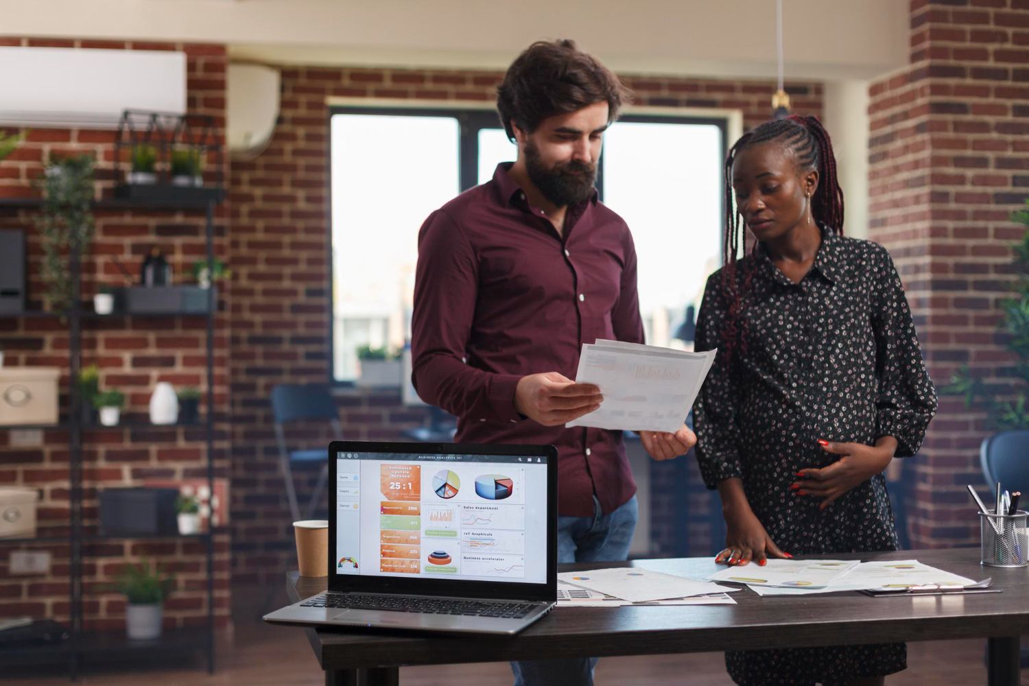 Two coworkers reviewing documents beside a laptop in a brick-walled office