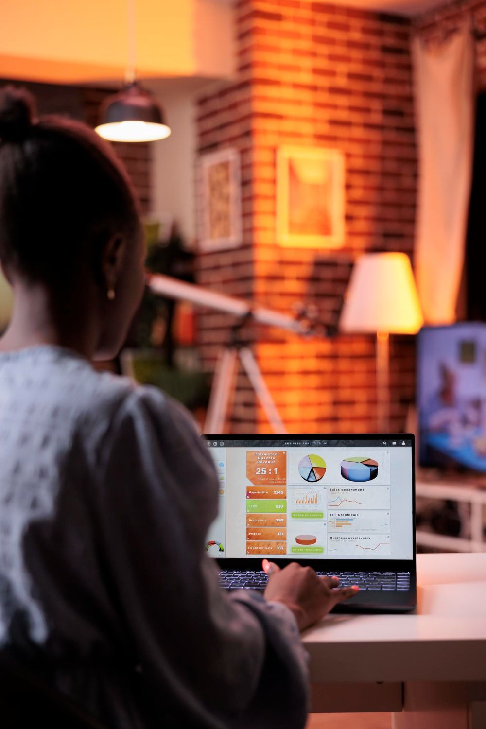 Person using a laptop at a desk in a cozy, warmly lit room with brick walls and a table lamp