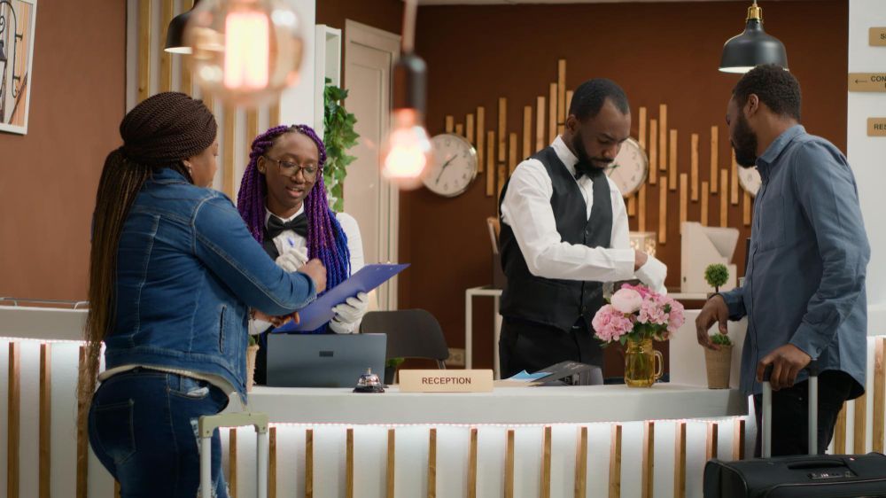 People checking in at a hotel front desk, with staff assisting and flowers on the counter.