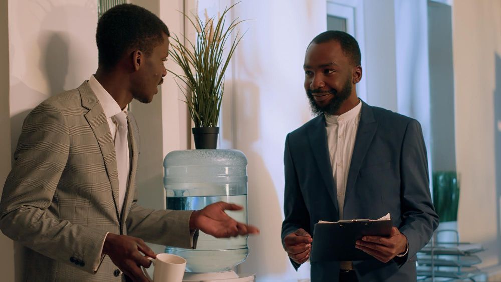 Two professionals talking in an office beside a water cooler, one holding a tablet and the other a cup