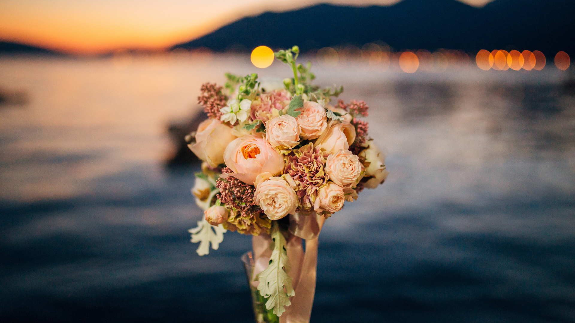 Bouquet of peach and white flowers in a glass vase, held against a blurred sunset waterfront backdrop.