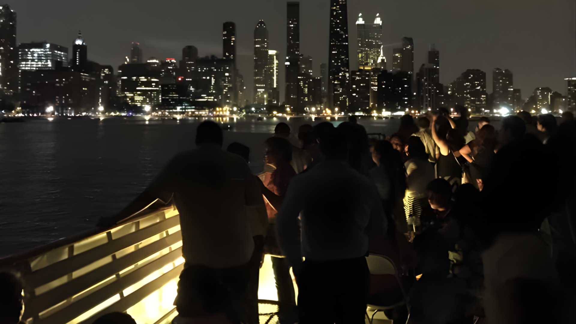People seated at glowing pink-lit tables on a dark rooftop or terrace at night, facing the water.