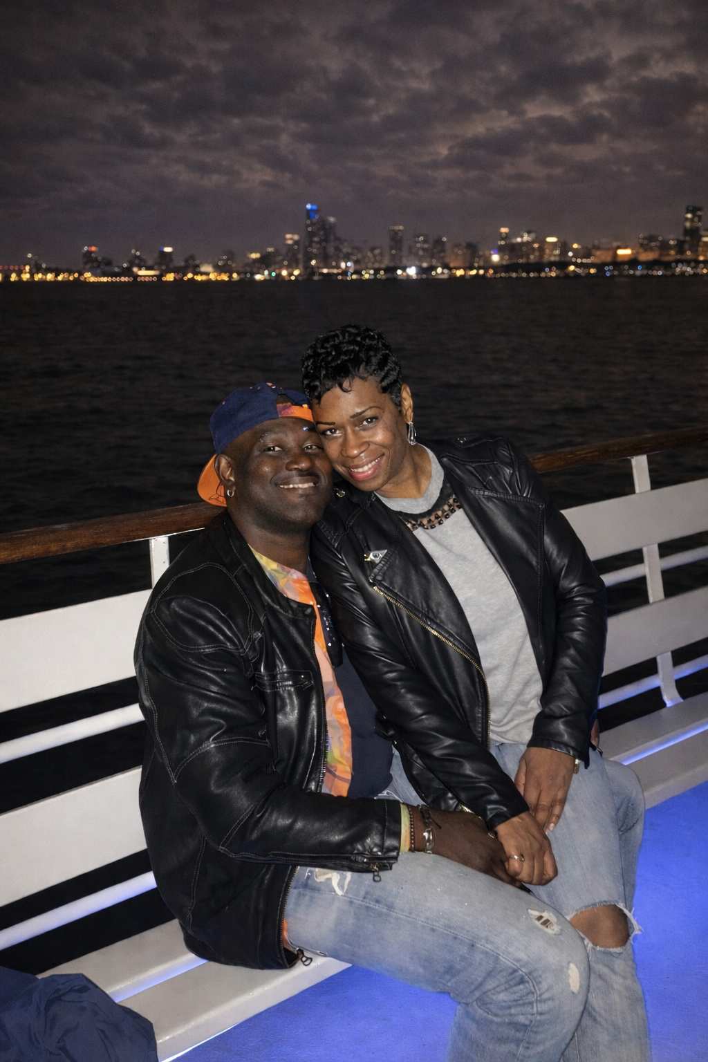 Two people smiling on a boat at night, with city lights and water in the background.