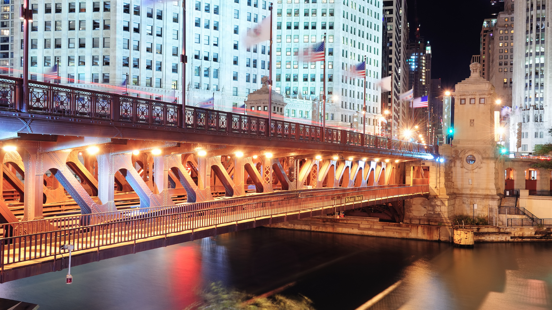 Nighttime city bridge lit in warm orange, spanning a river beside tall buildings.
