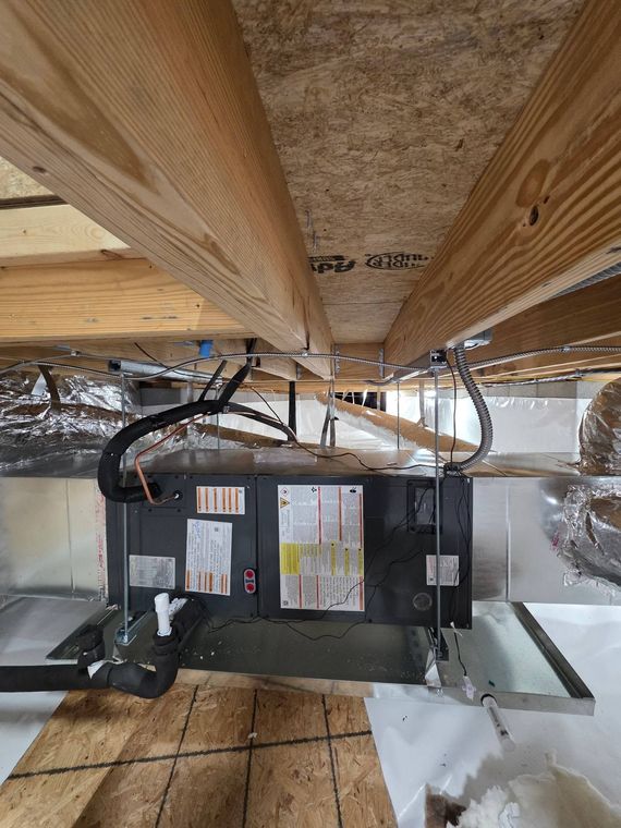 Furnace unit under wooden floor joists in a crawlspace, with ducting and wiring visible.