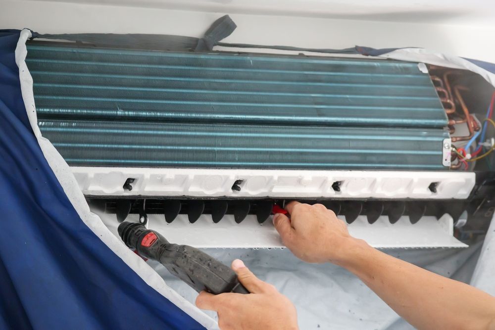 Hands working on a wall-mounted air conditioner with exposed internal parts and blue fins