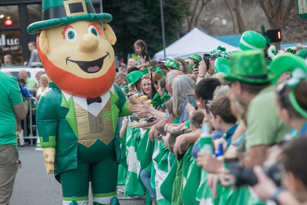 Leprechaun mascot in green suit at a St. Patrick's Day parade, waving to crowd wearing green.