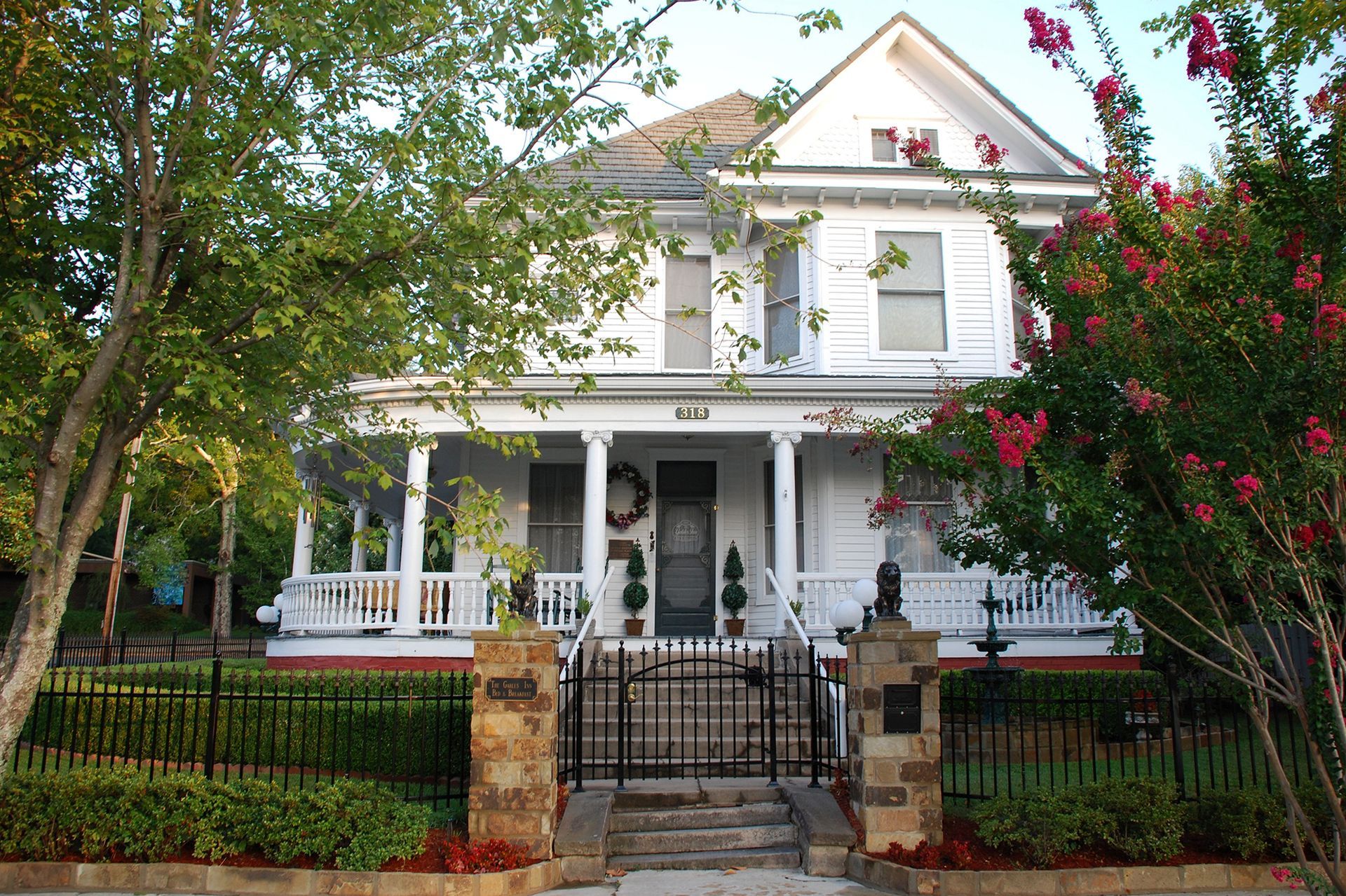 White two-story house with porch and black fence, framed by green trees and red flowers.