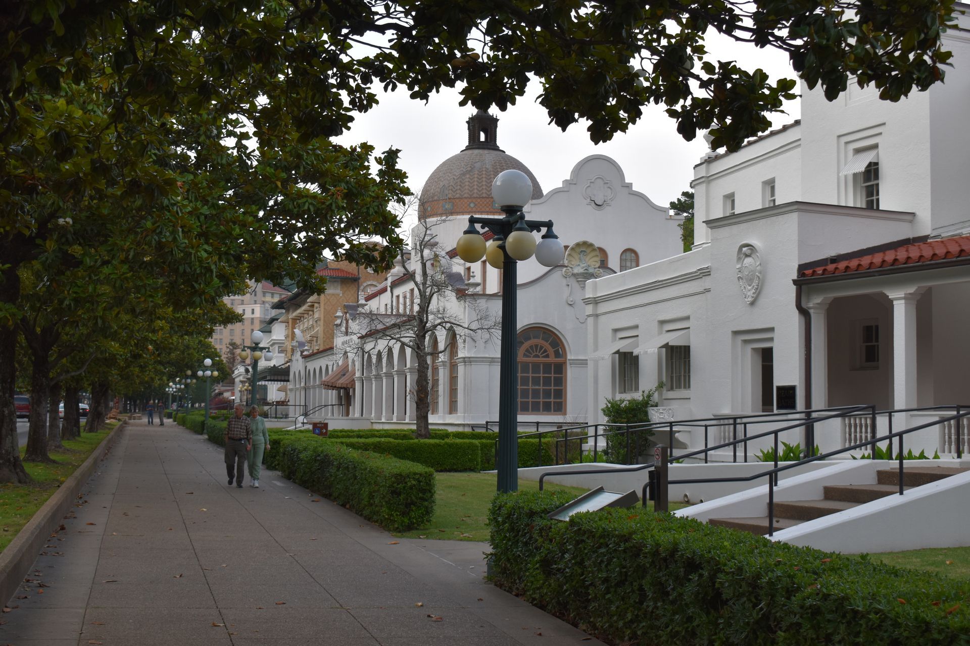 Sidewalk along park with white buildings, including a domed structure. People walk on the sidewalk, trees in the background.