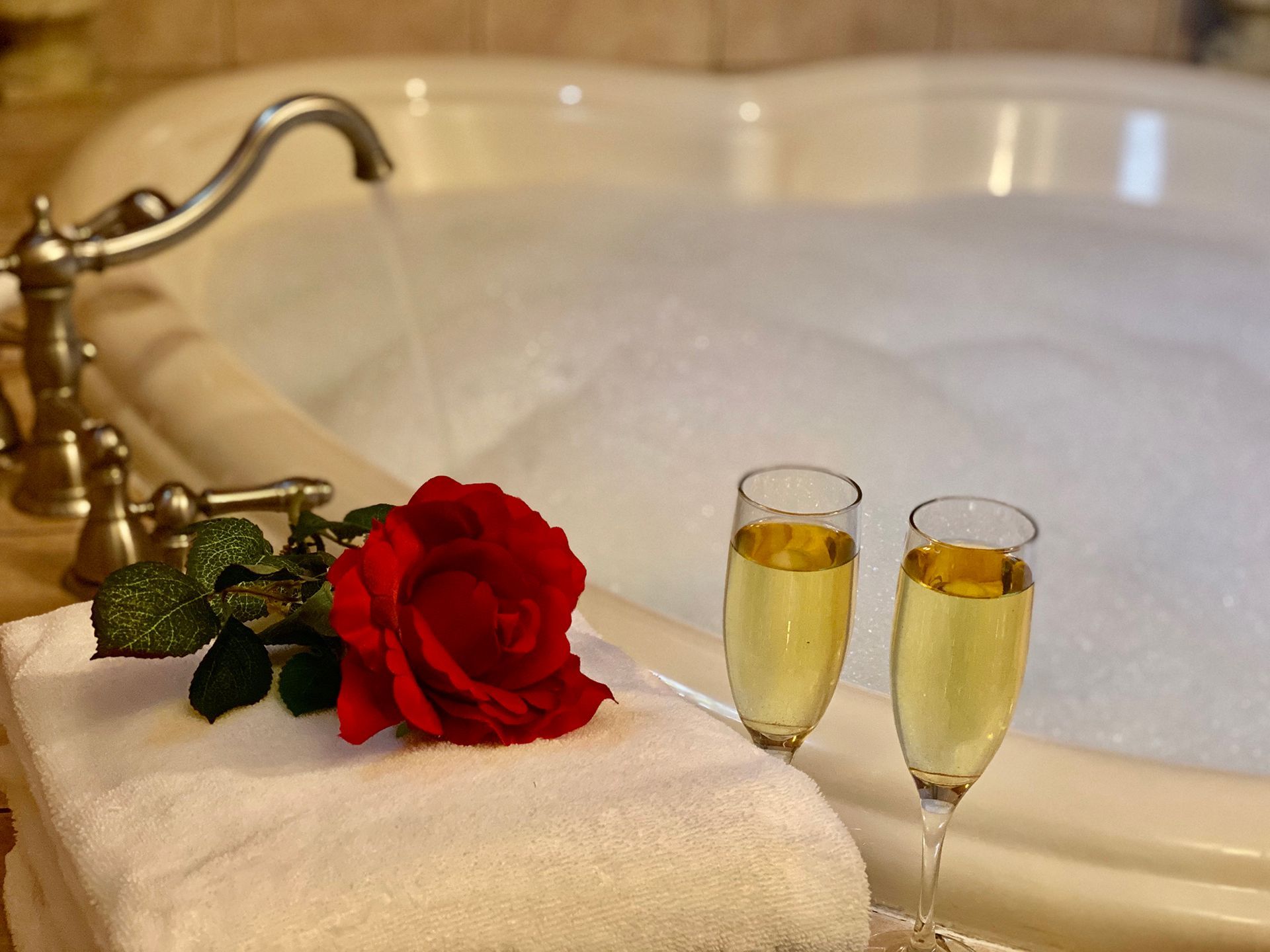 Champagne flutes, a rose, and towels near a heart-shaped bathtub filled with bubbles.