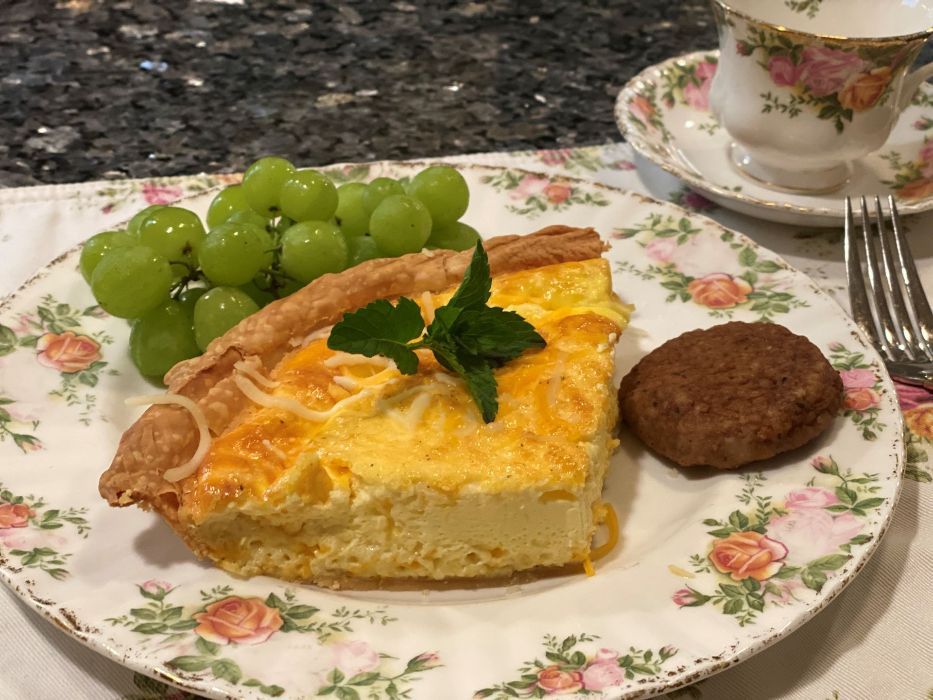 Slice of quiche, grapes, cookie, and teacup on floral plate; white table.