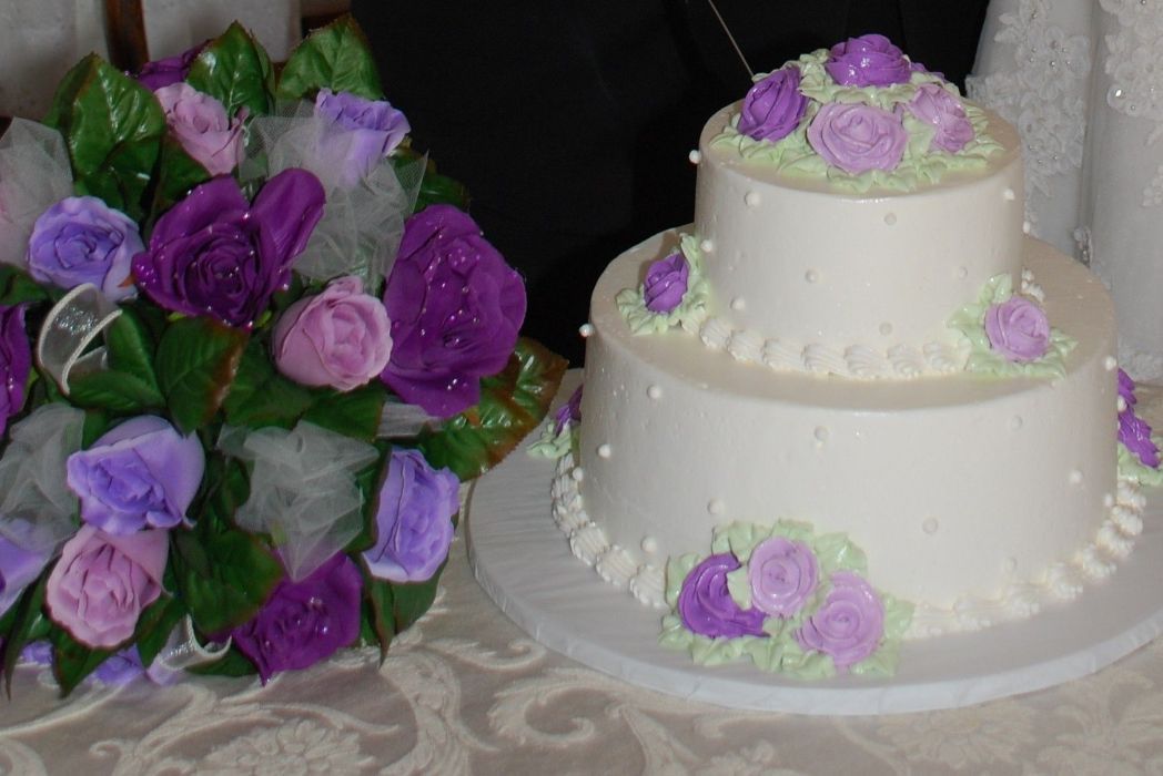 Two-tier white cake with purple floral accents, next to a bouquet of purple and pink roses.