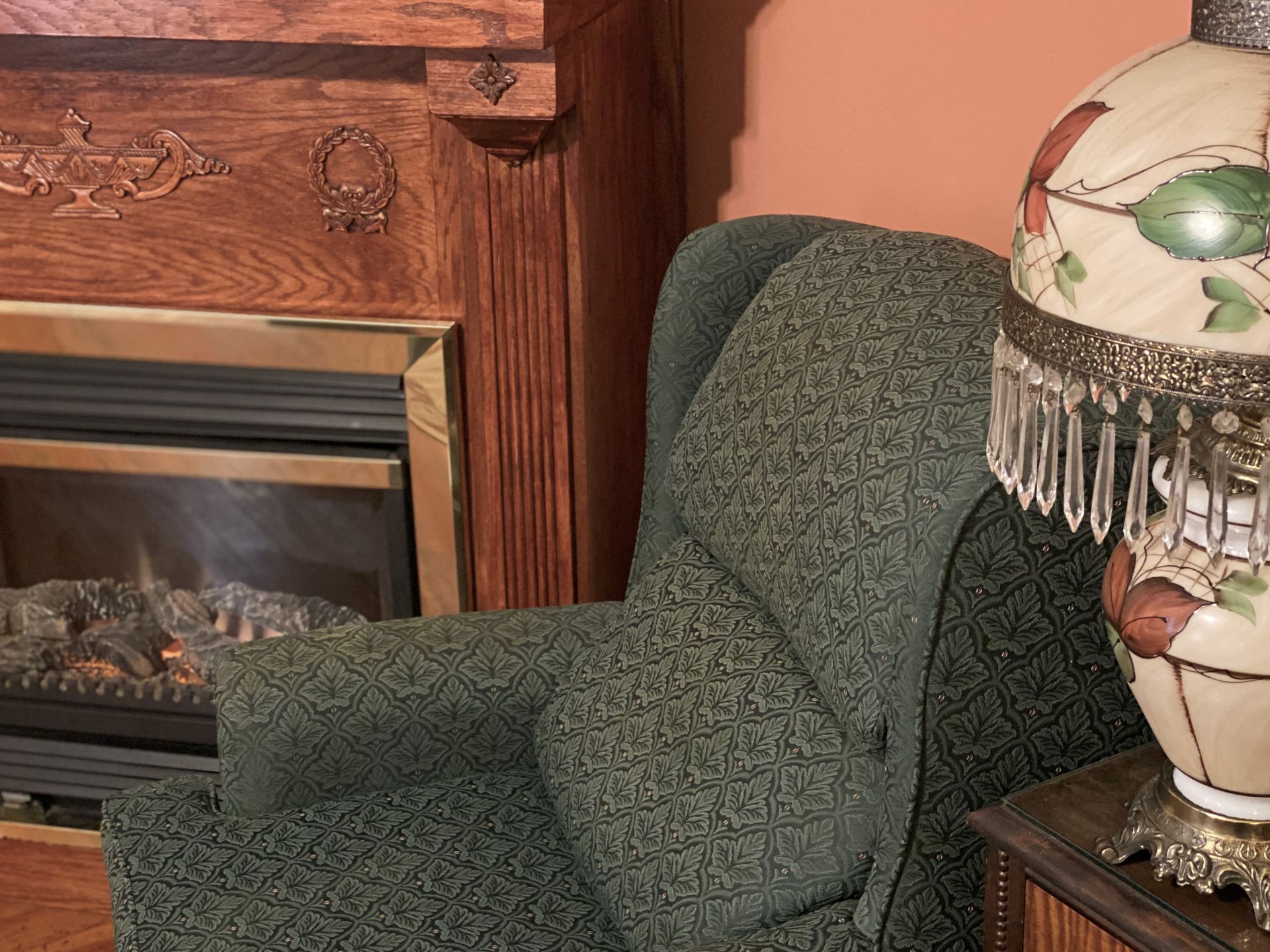 Cozy armchair next to lit lamp, fireplace with wooden trim in background.