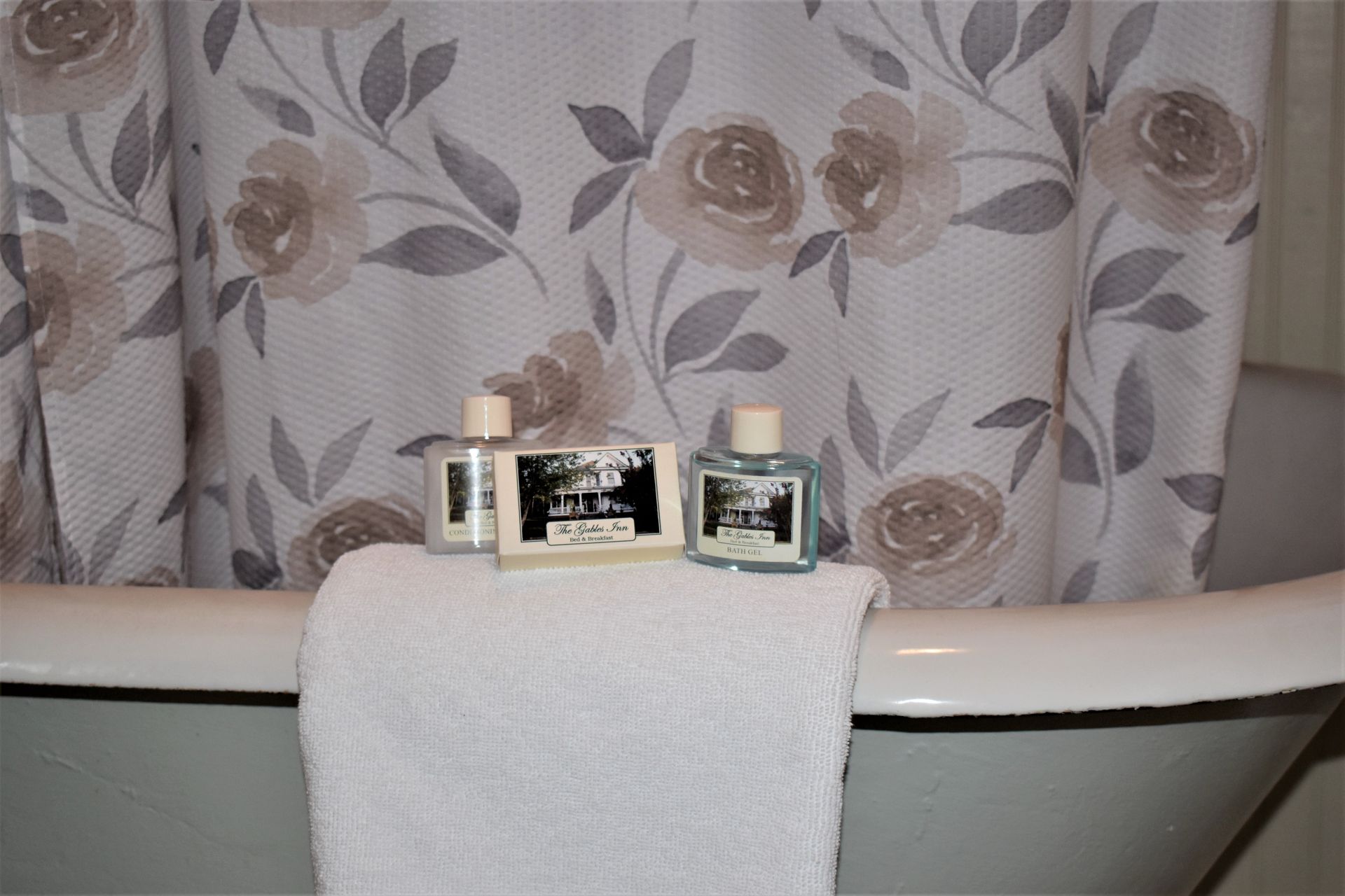 White towel on a bathtub rim with toiletries and floral shower curtain in the background.