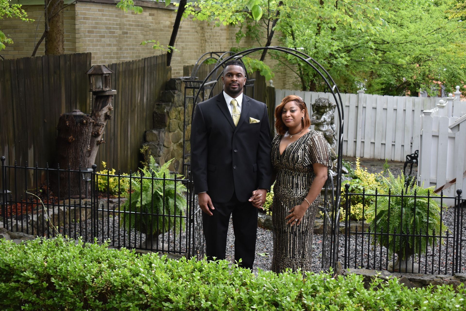 Couple in formal attire stands outside by greenery; man in black suit, woman in patterned dress.