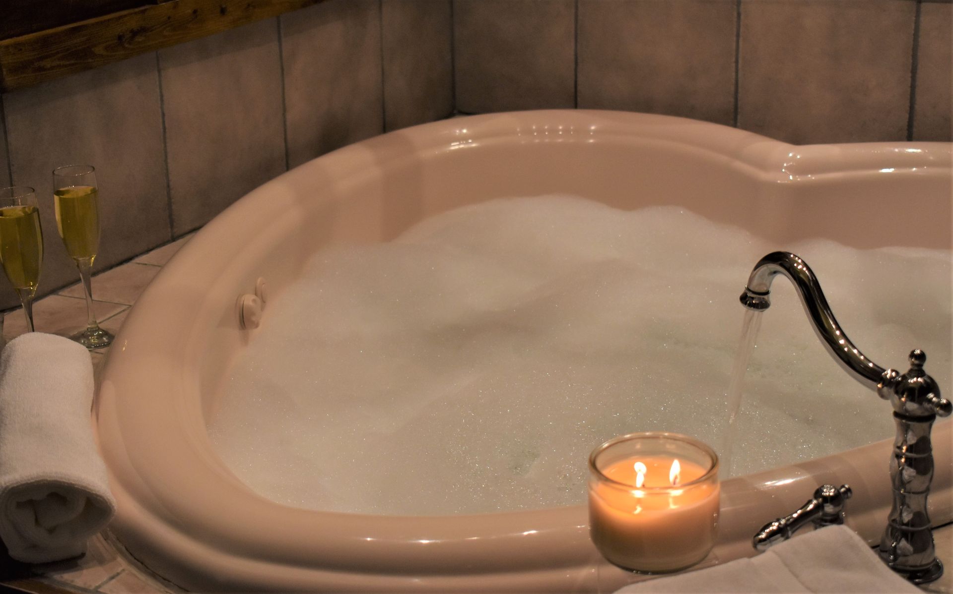 Heart-shaped bathtub filled with bubbles, candle lit, champagne glasses, and a rolled towel.