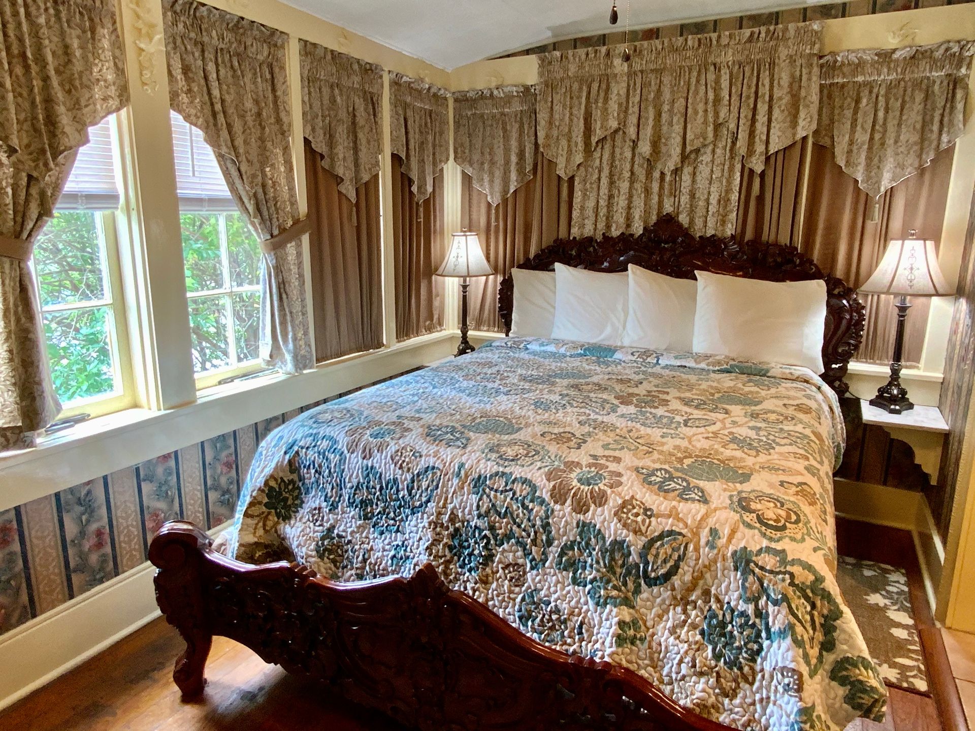 Bedroom with carved wooden bed, floral quilt, patterned wallpaper, and draped curtains.