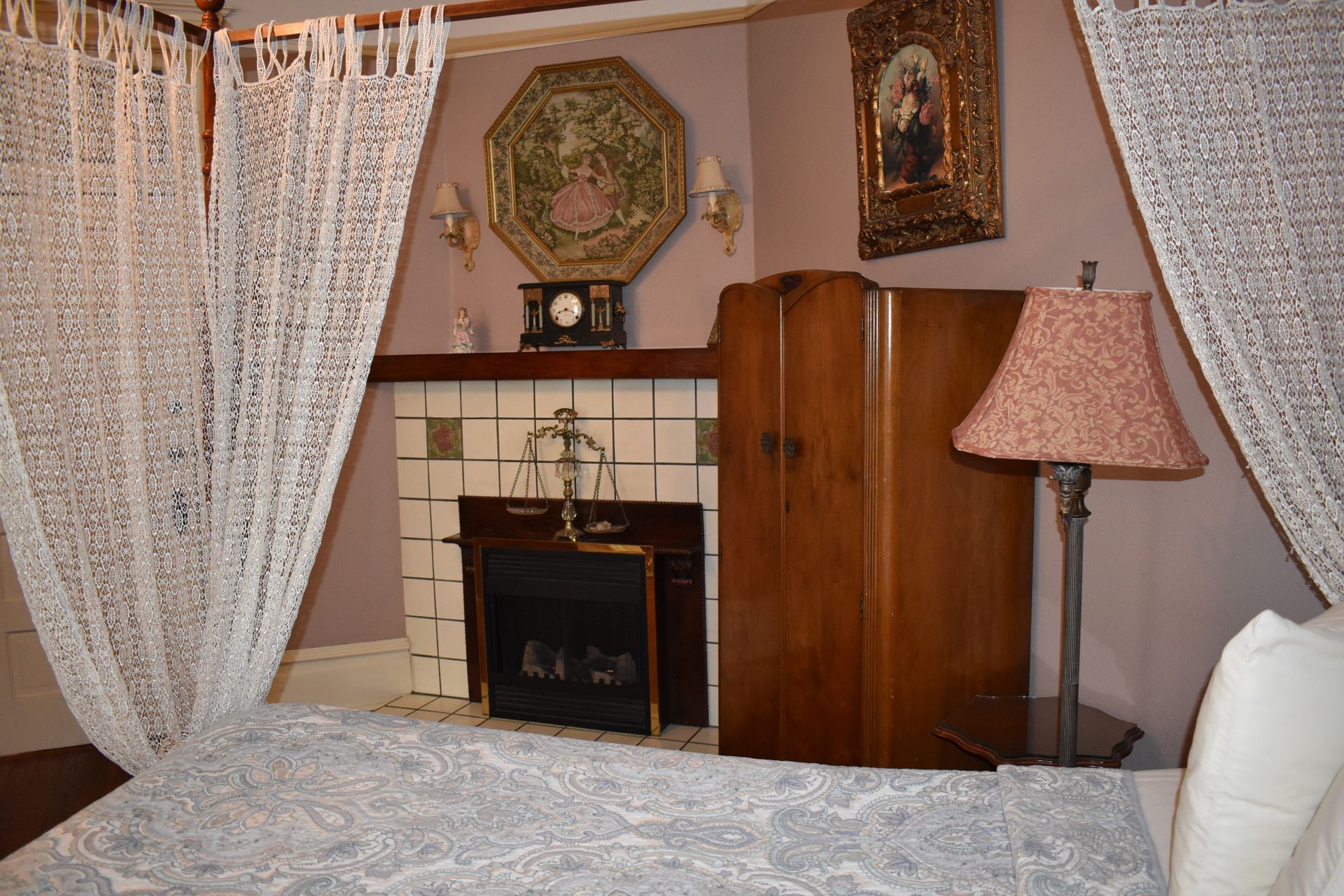 Cozy bedroom with a fireplace, decorative screen, and a lamp next to a bed framed by lace curtains.