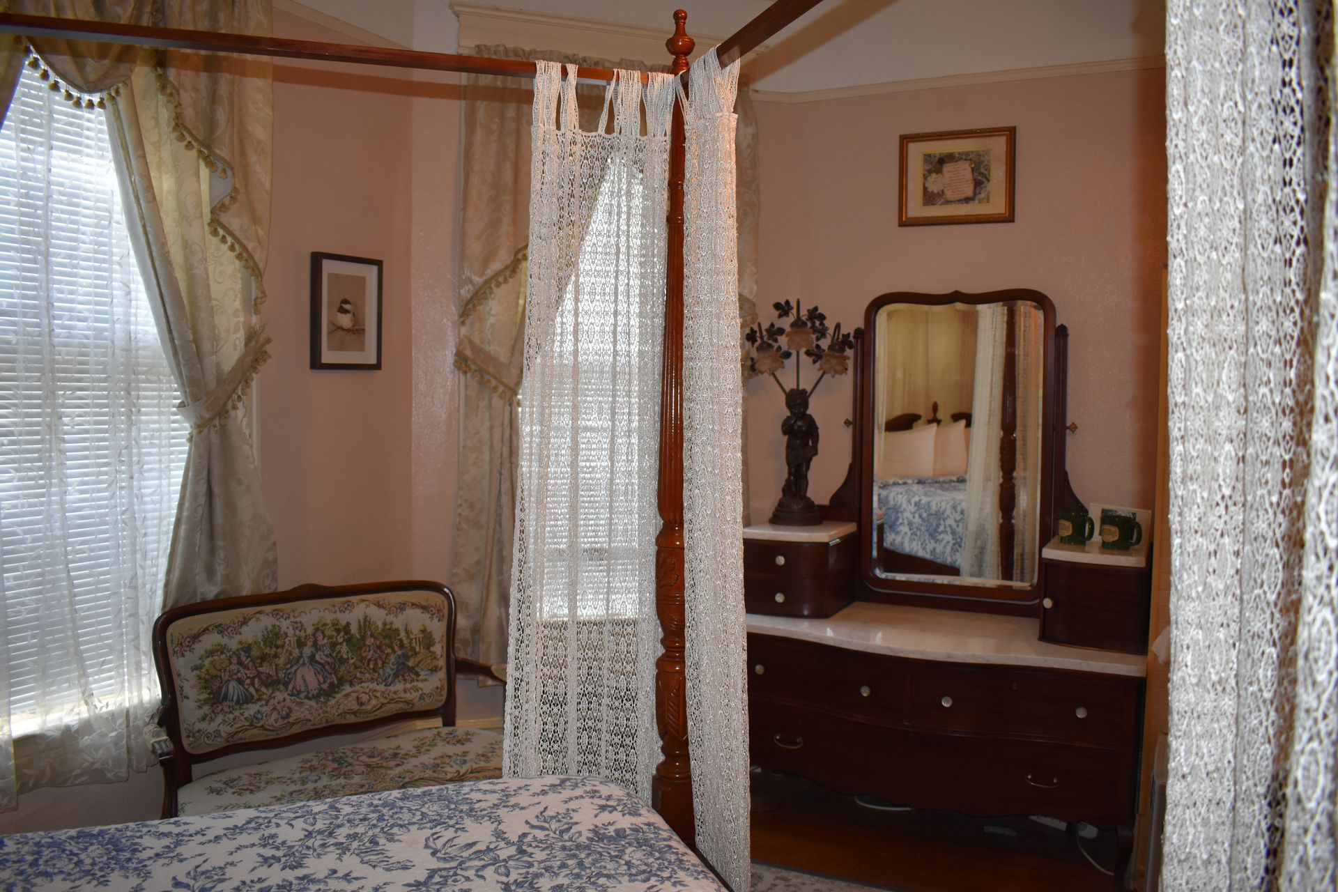 Bedroom with canopy bed, antique dresser, and chair; light pink walls and lace curtains.