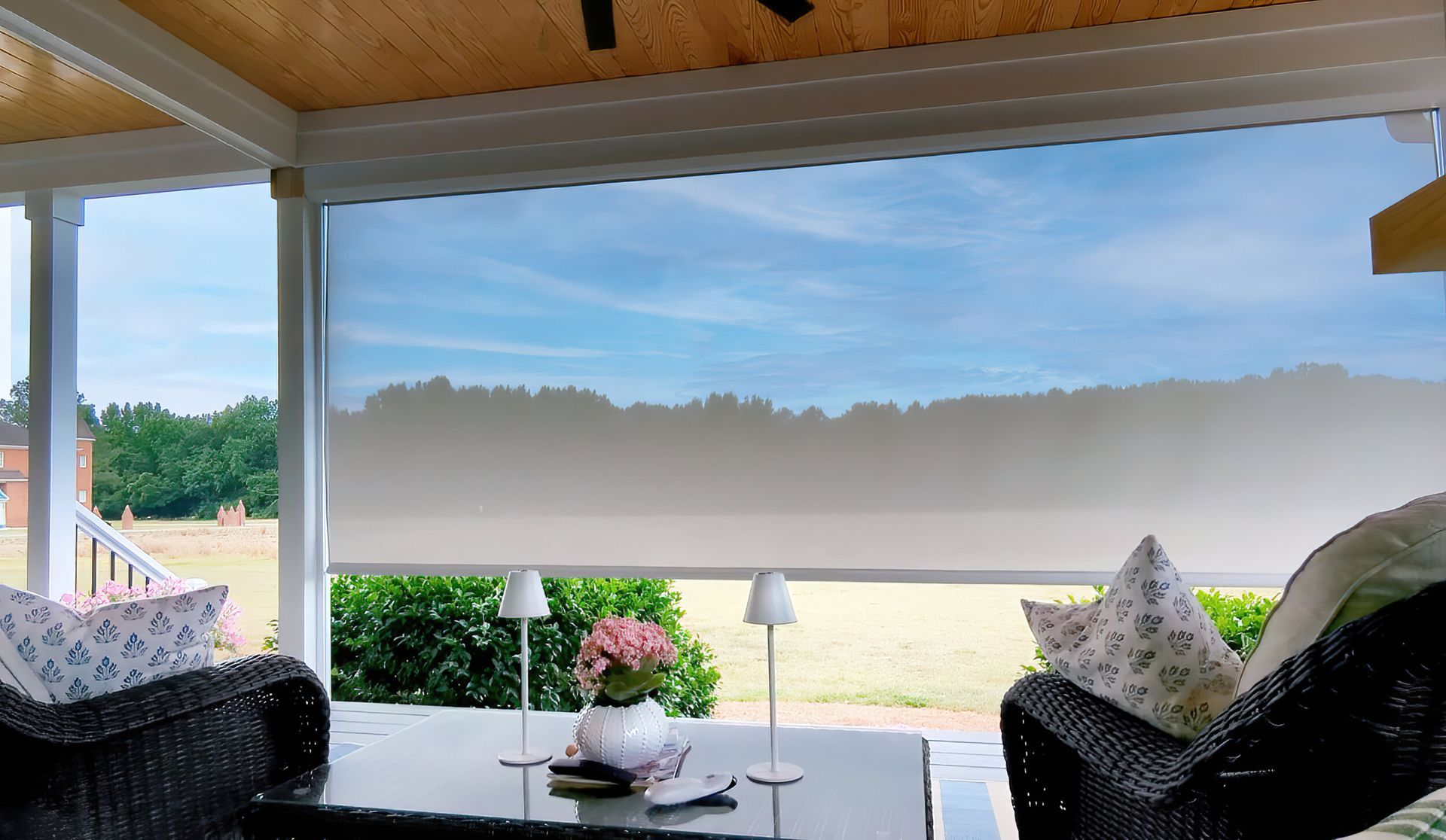 Covered porch with rolled-up shade; wicker furniture and tabletop decor overlook a sunny yard.