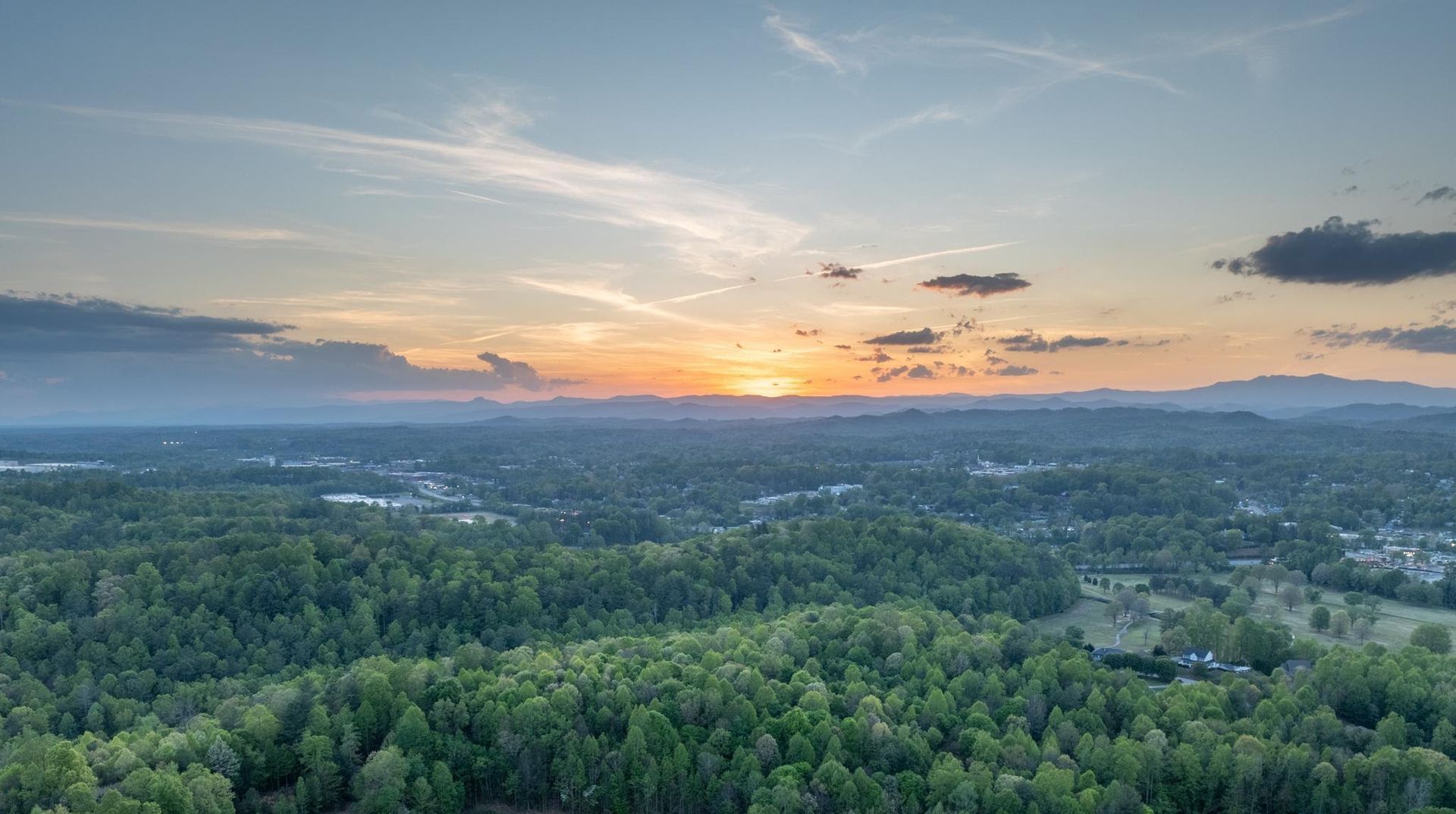 Sunset over a lush green forest with distant hills and a pastel sky