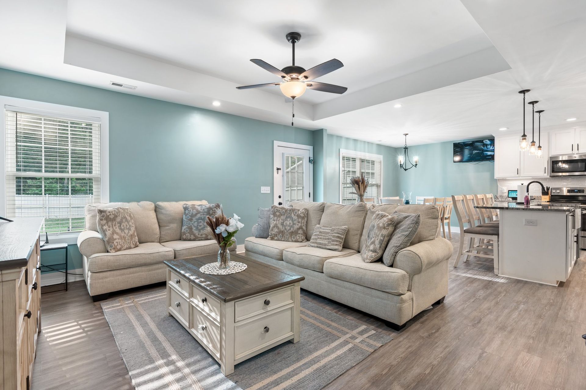 Bright open living room with gray sofas, white coffee table, ceiling fan, and kitchen island in the background