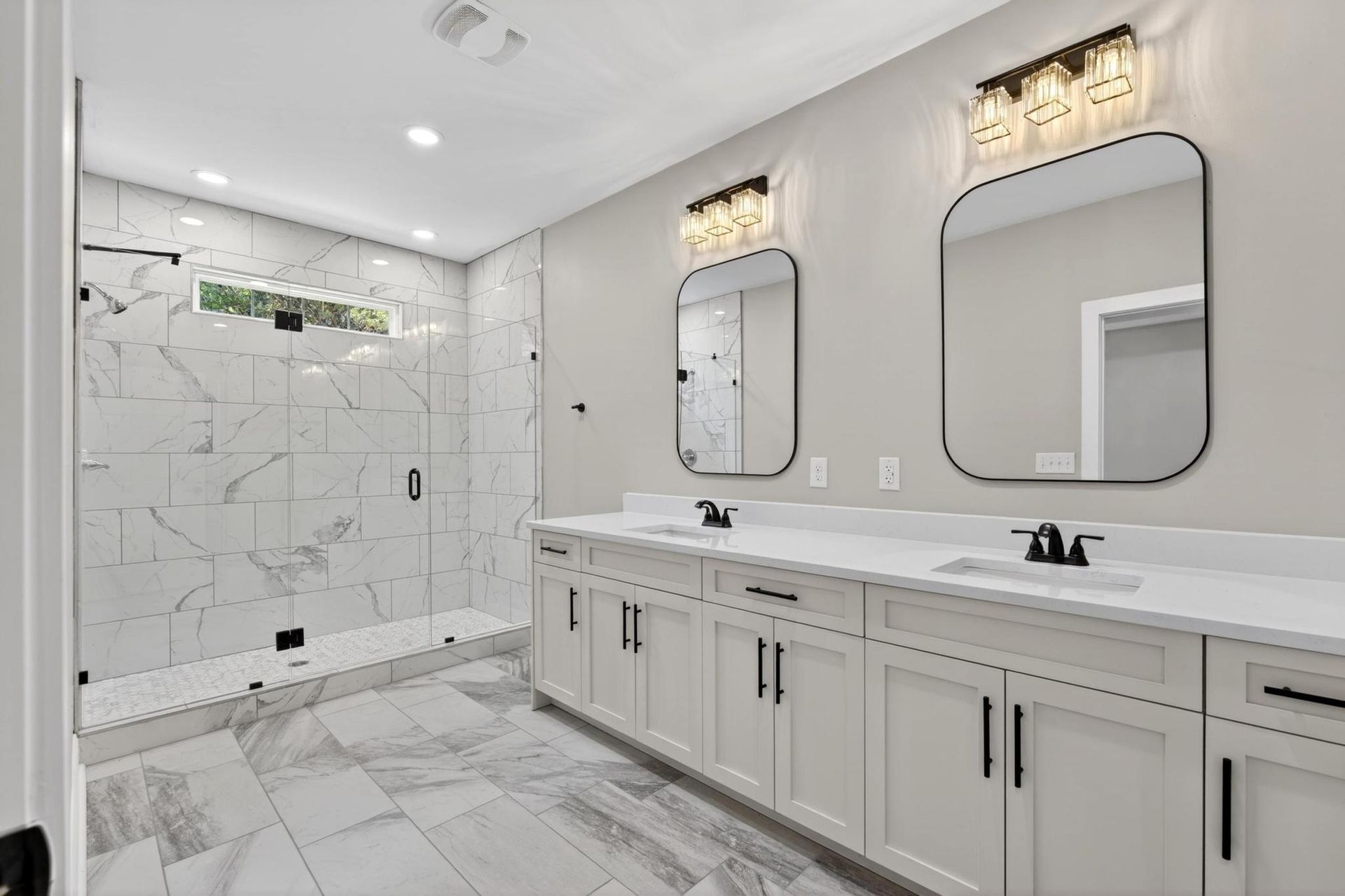 Bright modern bathroom with double vanity, large mirrors, marble tile, and a glass shower.