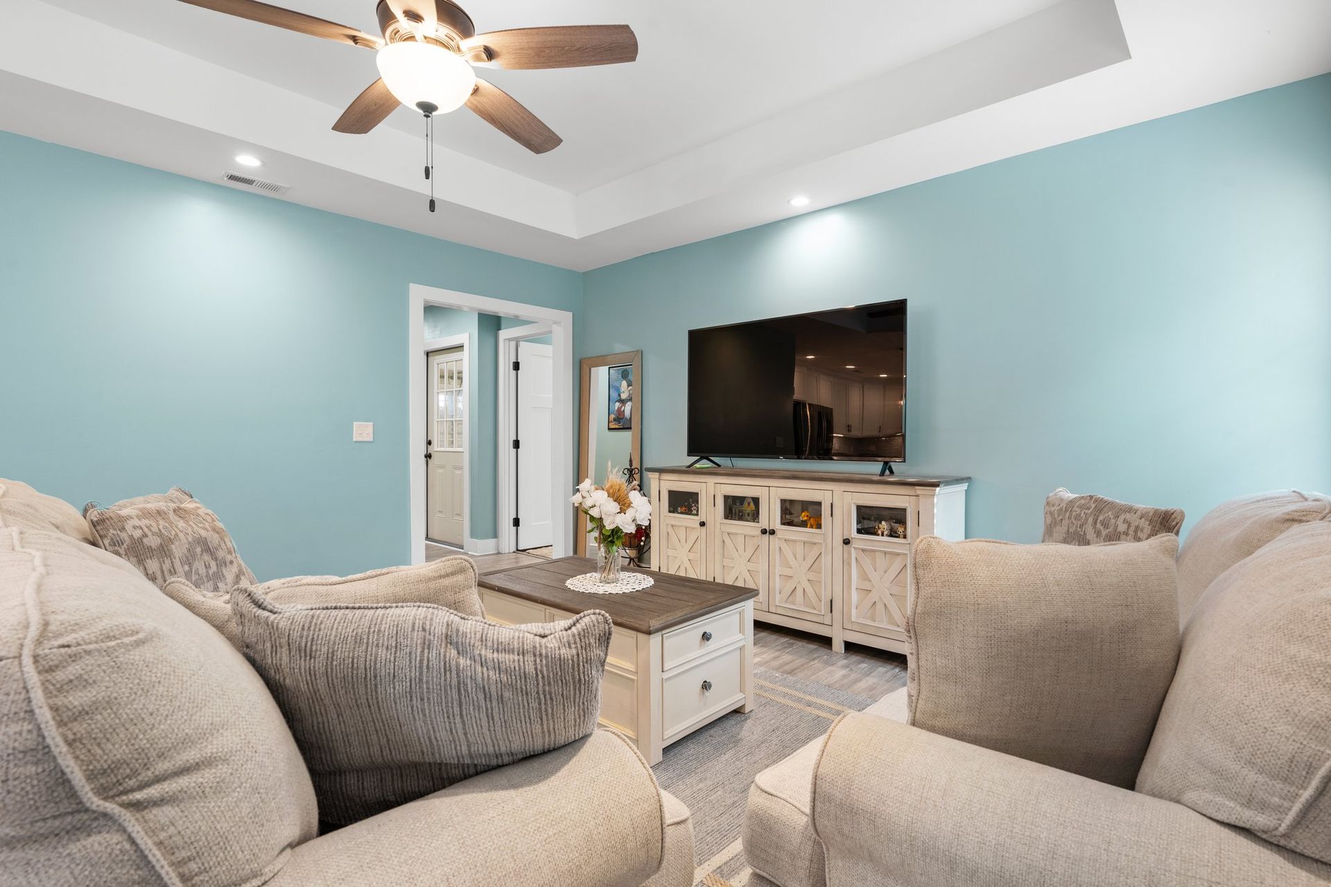 Bright living room with light blue walls, beige sofas, a coffee table, and a TV on a white console.