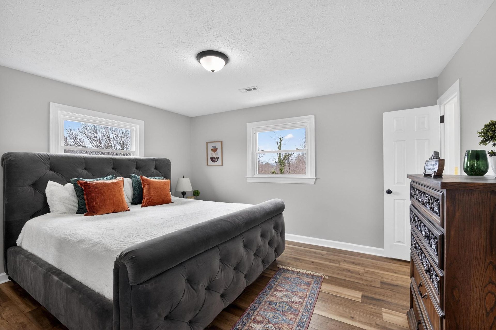 Modern bedroom with gray upholstered bed, patterned rug, wood dresser, and two windows.