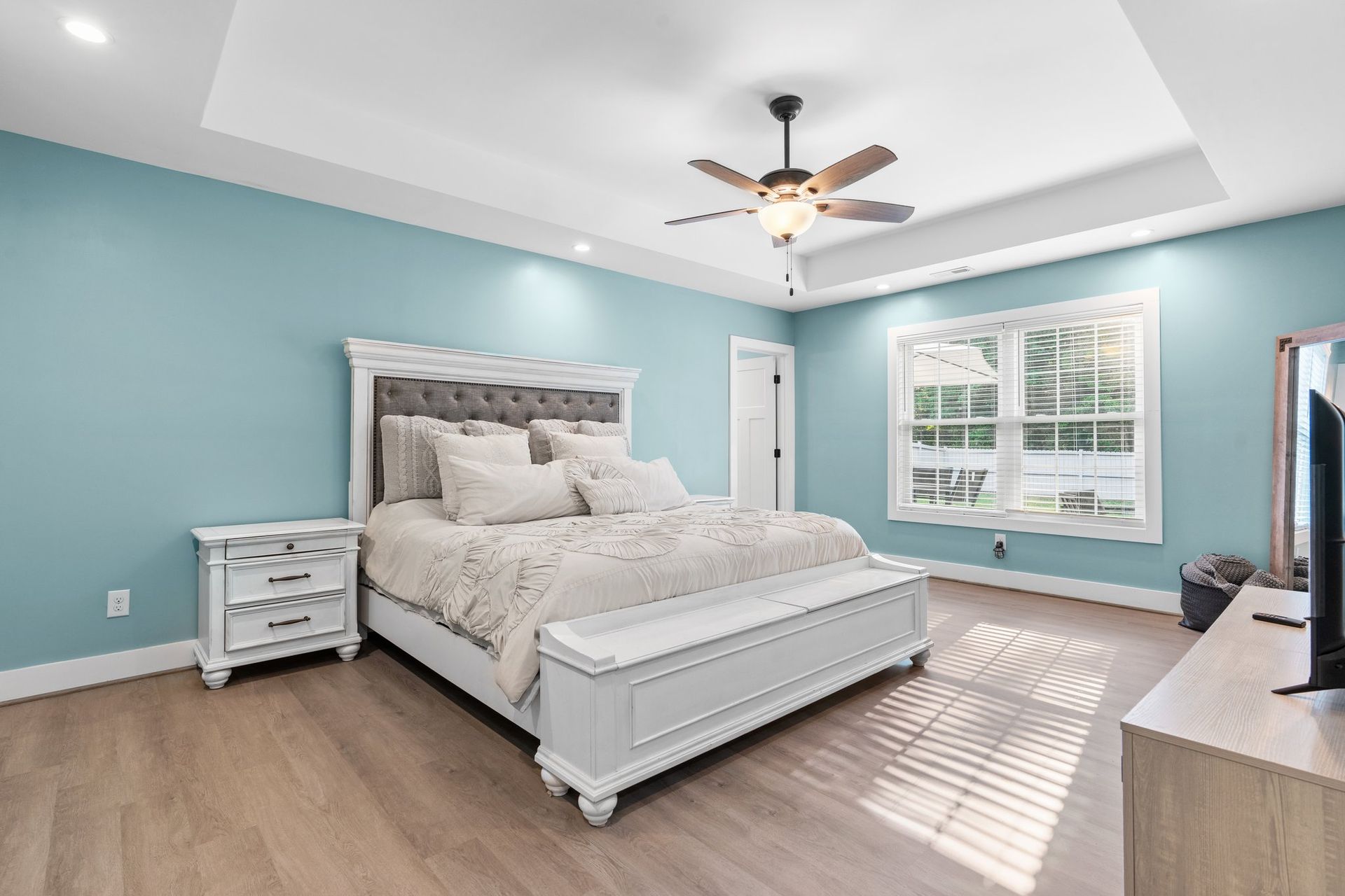 Bright bedroom with teal walls, white bed, ceiling fan, and sunlight from a large window.