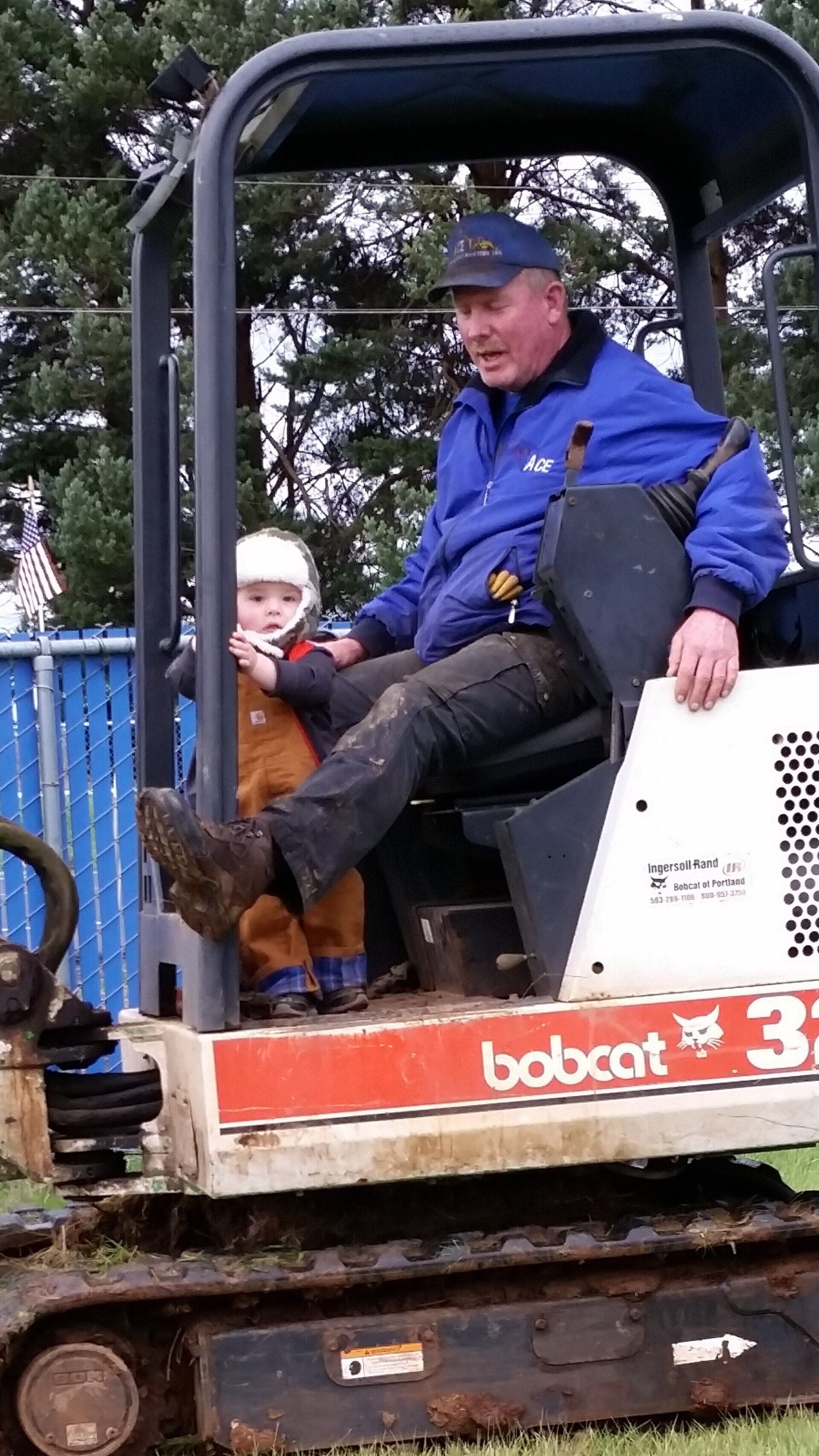 Man and child in a Bobcat excavator. Man in blue jacket smiles, child in overalls. Outdoors, cloudy day.