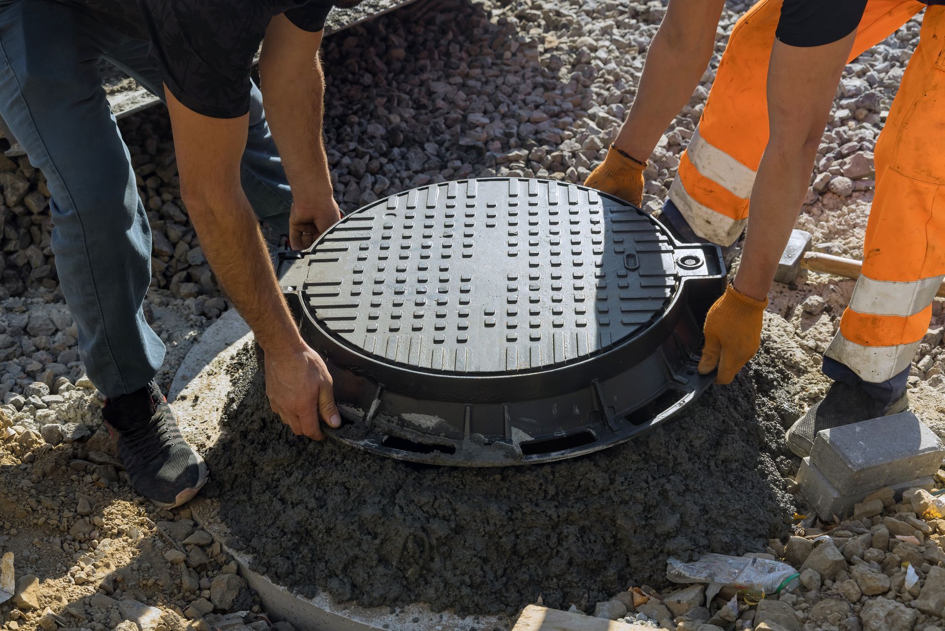 Two workers fitting a round black septic tank manhole cover onto a concrete base in gravel