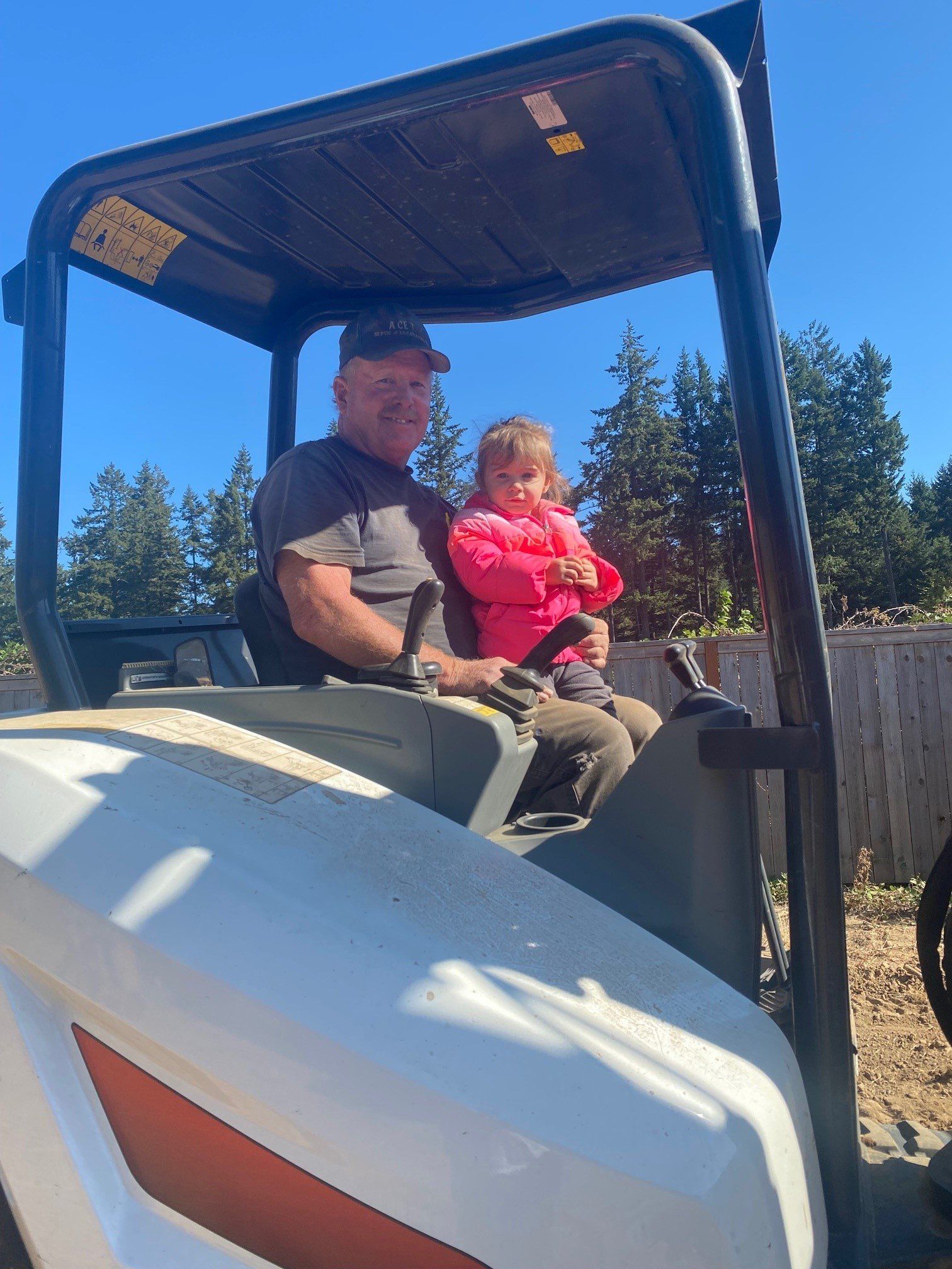 Man and young girl in a small tractor. They are outdoors on a sunny day. The girl is in pink
