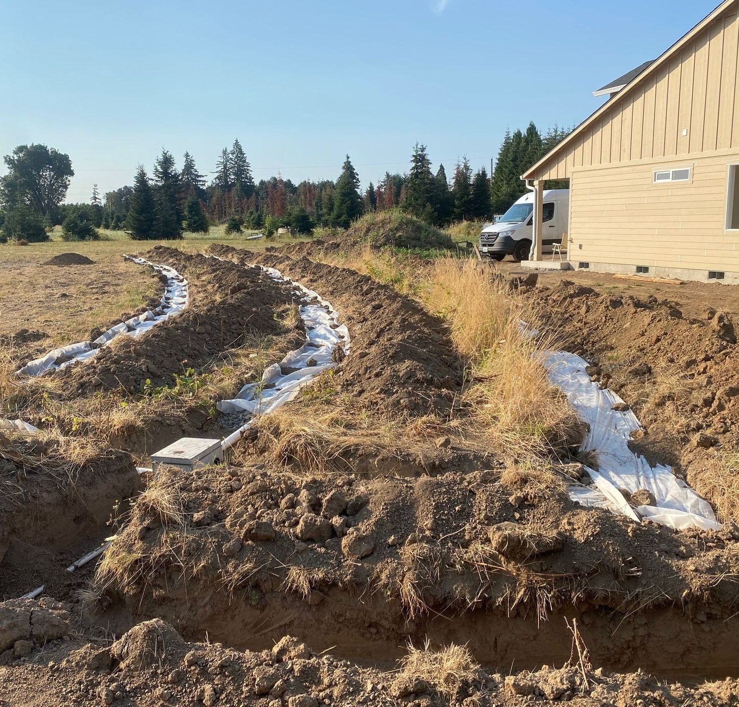 Trenches dug near a building site, revealing white septic system components. Dirt piles surround the work.