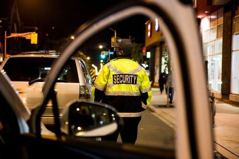 Security guard in a yellow jacket standing on a city street at night, seen through a car window.