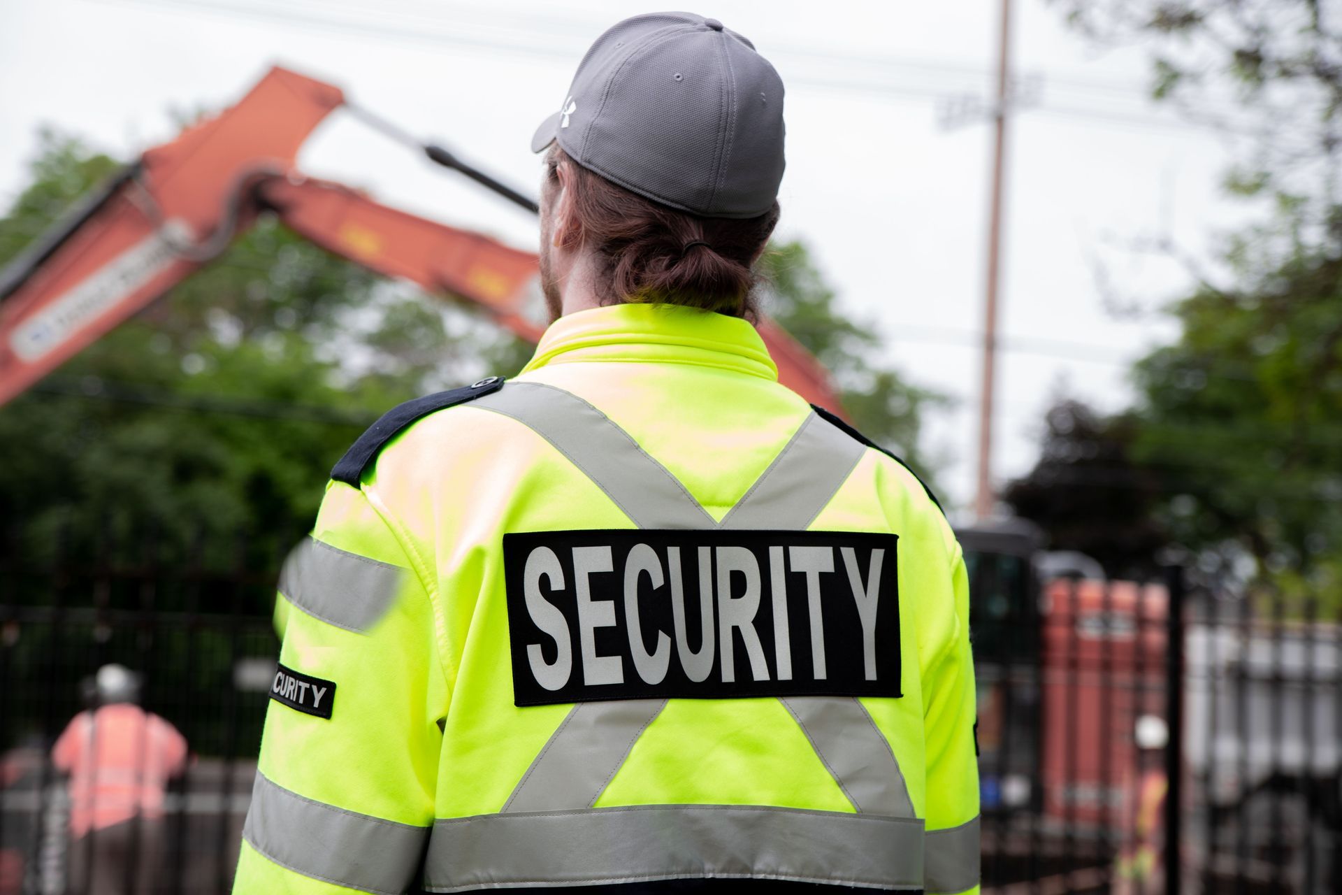 Security guard in neon yellow jacket with reflective stripes, seen from behind outdoors near a fenced area.
