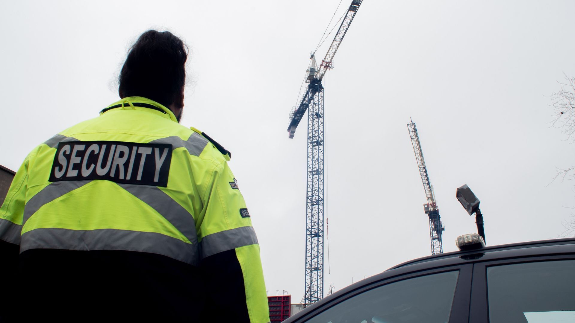 Security guard in neon jacket watching construction cranes beside a parked car.