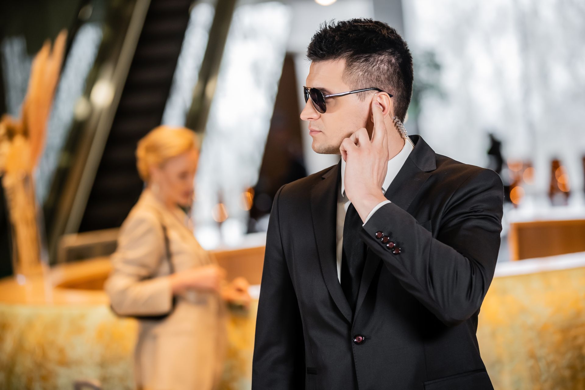 Man in a black suit and sunglasses using a phone outdoors, with a blurred woman in the background