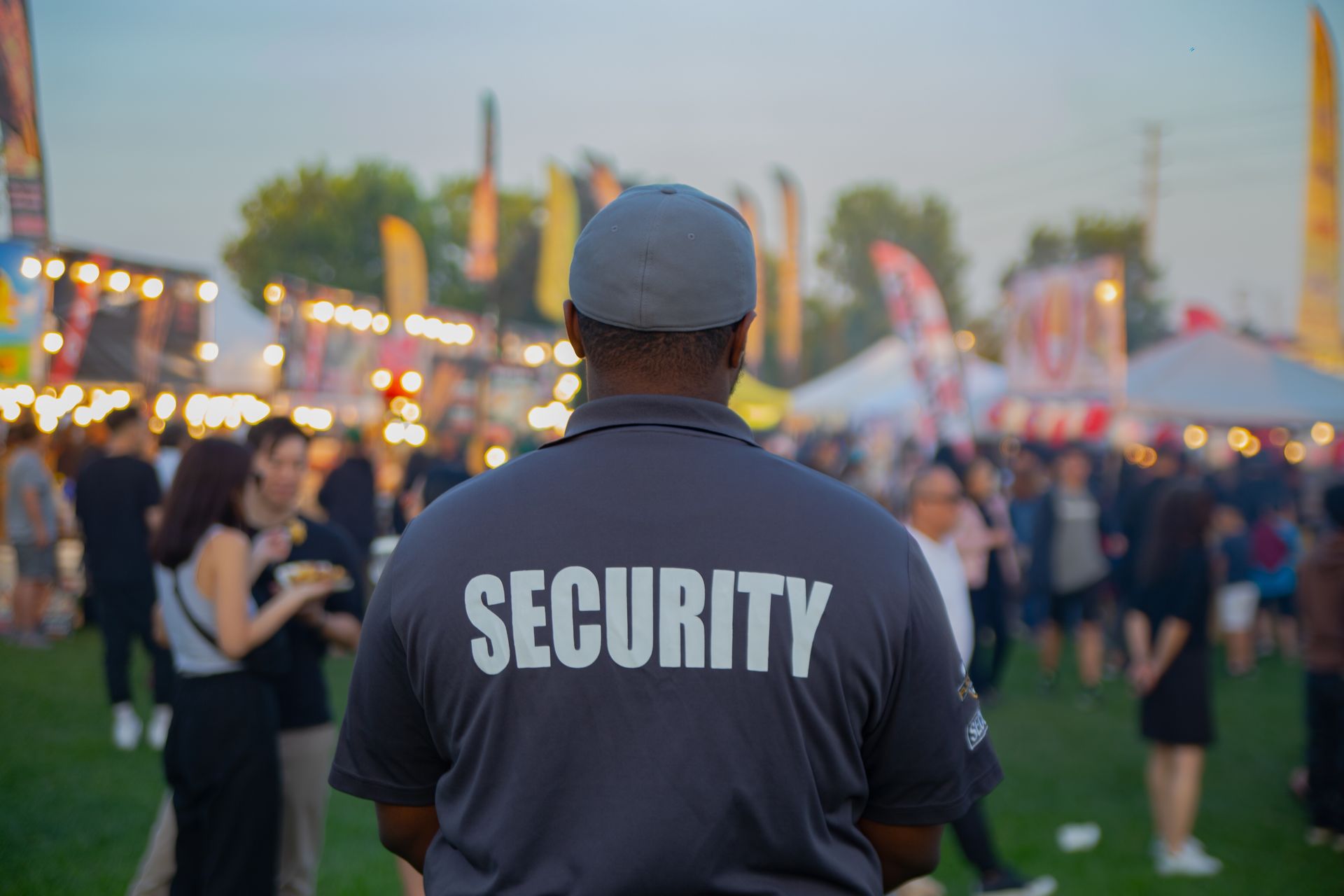 Security guard in navy shirt at an outdoor event, facing a crowd at dusk