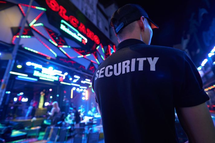 Security guard standing in a neon-lit arcade hallway, seen from behind