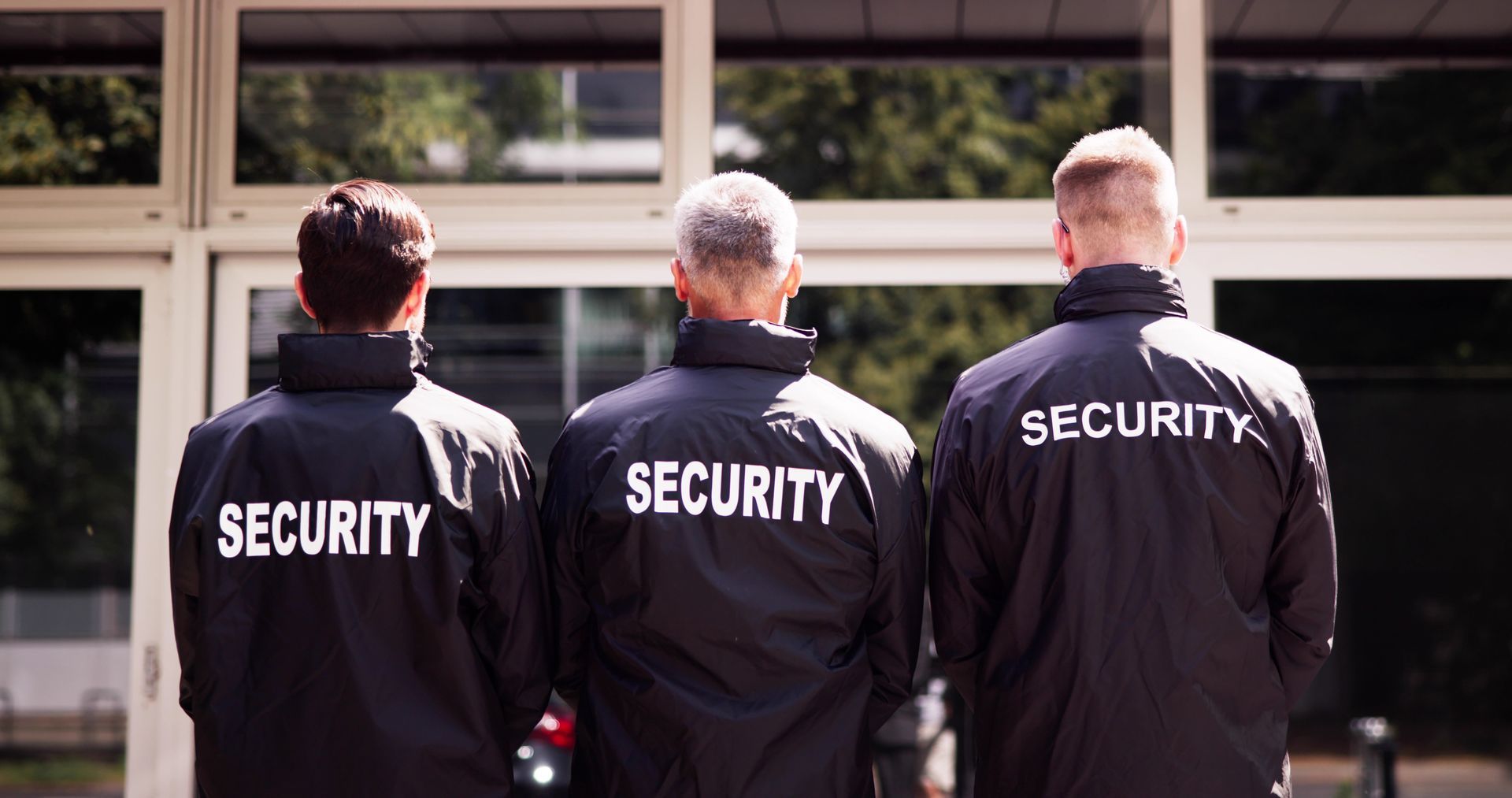 Three security guards in black jackets stand with their backs to the camera outside a building.